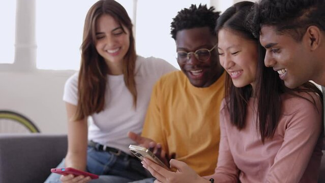 Young group of diverse friends sitting on sofa using mobile phone. Smiling multiracial teenage people laughing having fun together watching funny social media video content on smart phone at home
