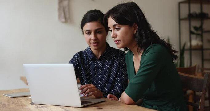 Two multi ethnic women colleagues staring at laptop screen, learn new task or discuss corporate software, prepare presentation, search solution, mentor explaining program. Workflow use tech, teamwork
