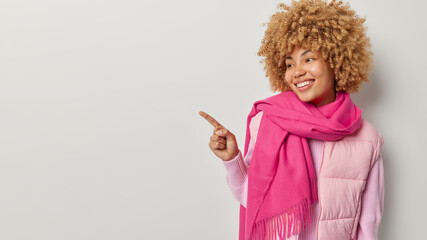 Fashionable curly haired woman in vest and pink warm scarf points index finger left shows promo offer demonstrates banner smiles gladfully isolated over grey background. Follow this direction