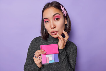 Portrait of young Asian woman with hairpin holds colorful eyeshadow palette wears vivid makeup keeps hand on face dressed in black turtleneck isolated over purple background. Beauty concept.