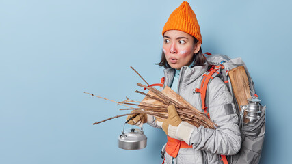 Shocked embarrassed young Asian female traveler or hiker with backpack holds kettle and wood looks...
