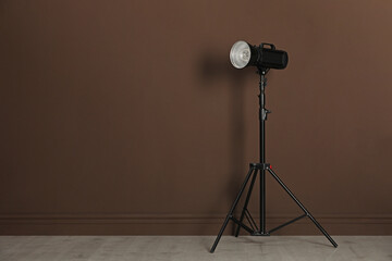 Studio flash light with reflector on tripod near brown wall in room, space for text. Professional photographer's equipment