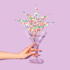 Martini glass with colorfool glitter and a woman's hand on pastel purple backgraund . Party drink...