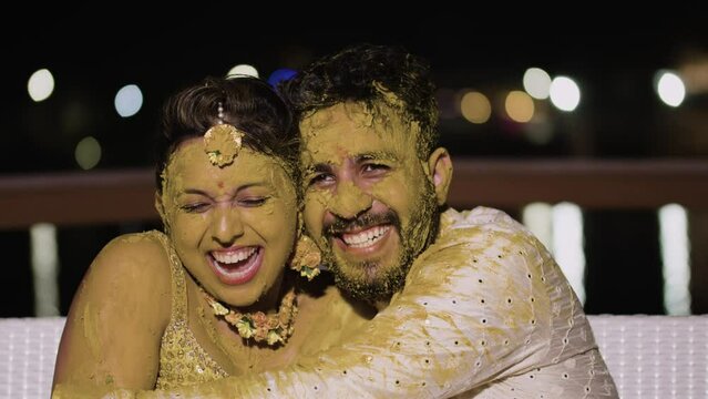 Happy Indian Groom Hugging His Beautiful Bride With Turmeric Applied On Their Faces At Haldi Function During Their Wedding Week. - Medium Closeup Shot