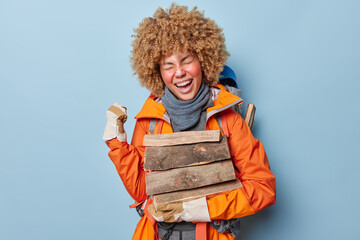 People and camping concept. Positive curly haired female camper poses with pile of wood clenches fist feels very happy wears orange jacket and gloves has active rest in nature isolated on blue wall