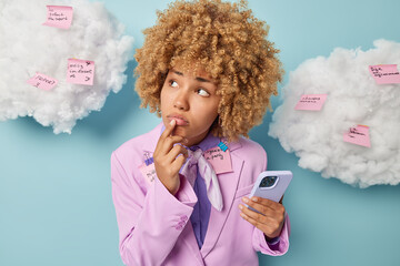 Upset female student or office worker looks sadly somewhere being deep in thoughts dressed formally holds mobile phone tries to remember something uses memo stickers to write list to do. Too much work