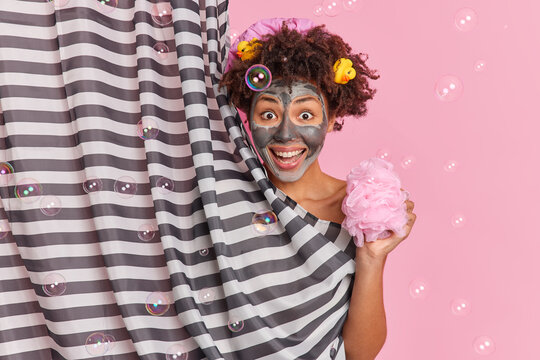 Positive Curly Haired Woman With Rubber Ducks In Hair Applies Beauty Wellness Mask Holds Sponge Takes Shower Hides Behind Striped Curtain Isolated Over Pink Background With Soap Bubbles Around