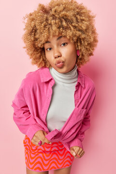 Vertical shot of beautiful romantic woman keeps lips folded wears fashionable clothes sends mwah at camera looks directly at camera isolated over pink background. Facial expressions concept.