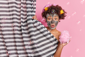 Skin care hygiene and beauty rituals concept. Shocked curly haired woman applies facial mask for reducing blackheads takes shower in bathroom holds sponge for washing body soap bubbles around