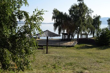 On the shore of the lake there is a beach umbrella, sun loungers, sand, lifebuoy, lawn and birch trees on a summer morning