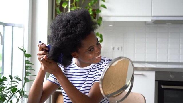 Young happy African American teenage girl grooming herself doing traditional afro hairstyle. Smiling woman in striped T-shirt brushes hair and looks in mirror sits in kitchen of own apartment