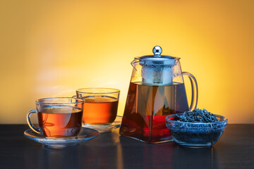 Glass teapot and two glass cups with tea on dark table.