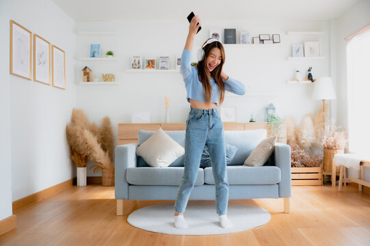 Young Asian Woman Dancing In Living Room. Asian Woman With A Smile Relaxing At Home, Happy And In Good Mental And Physical Health, Wearing Headset And Listening To Music