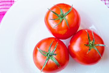 Three red ripe tomatoes with green stems and water drops on a white plate-top view