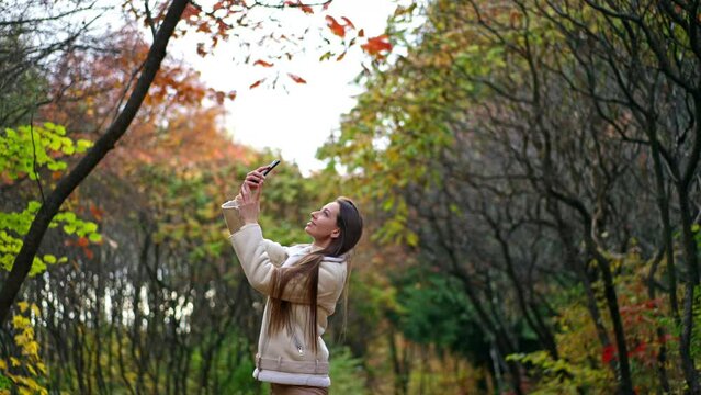 Young attractive long-haired lady taking picture of nature on her phone. Smiling positive woman enjoying beautiful colorful leaves in autumn.