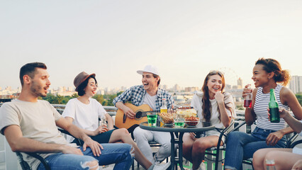 Handsome young man is playing the guitar while his cheerful friends are singing, drinking and laughing sitting at table on rooftop. Music, fun and summertime concept.