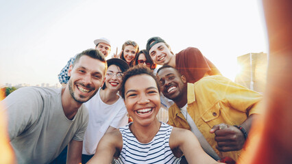 Point of view shot of pretty African American girl holding camera and taking selfie with happy friends at party on roof. Men and women are looking at camera, posing and laughing. - 542324677