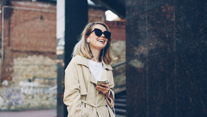 Pretty girl is listening to music through earphones using smartphone and dancing walking in the street and enjoying rhythm and modern city. Girl is wearing sunglasses and trendy coat.