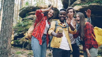 Fototapeta na wymiar Happy African American guy is making video call using modern smartphone during hike in forest with friends, young people are looking at screen, waving hand and talking.