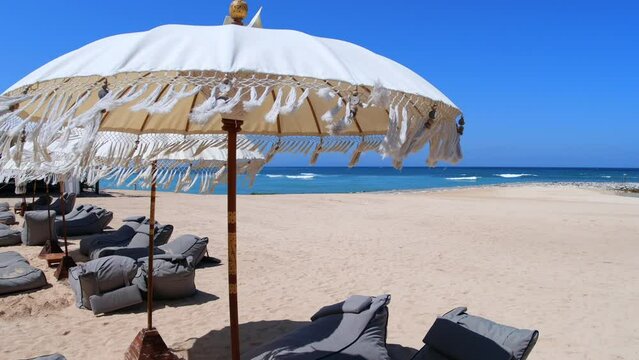 Along the seashore on a white sand beach, beach chairs with umbrellas stand alone, waiting for their visitors. Umbrellas create a pleasant shade from the hot sun.