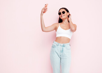 Young beautiful smiling female in trendy summer clothes. Sexy carefree woman posing near pink wall in studio. Positive brunette model looking at cellphone screen. Taking selfie photos