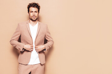 Portrait of handsome confident stylish hipster lambersexual model. Sexy modern man dressed in elegant suit. Fashion male with curly hairstyle posing in studio. Isolated on beige