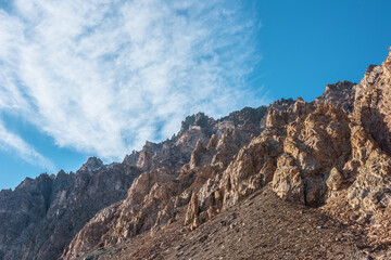 Scenic mountain landscape with sharp rocks under cirrus clouds in sunny day. Colorful scenery with gold sunlit sharp rocky mountains. High rocky mountains in golden sunlight under spindrift clouds.