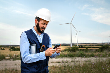 Happy male electric engineer wearing helmet and vest checking mobile phone in wind turbine farm.