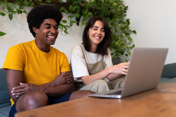 Smiling young multiracial couple sit on the couch together using laptop to chat with family in a video call.