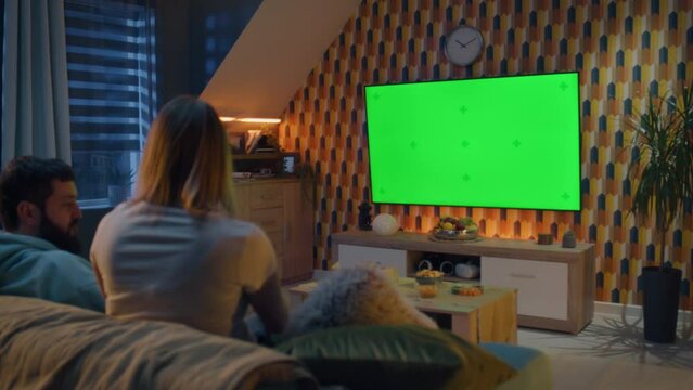 Woman jumping on couch. Couple on sofa in living room, watching breaking news, movie or series on TV with snacks. Wife and husband resting at home in the evening together. Green screen. Chromakey.