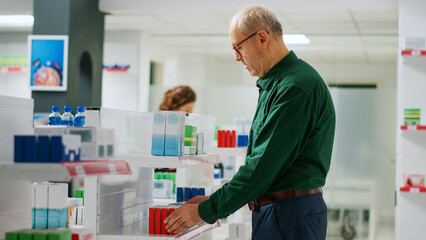 Senior client looking at medicaments boxes in pharmacy, buying pharmaceutical products from drugstore shelves. Elderly man checking packages and leaflet of pills and capsules, having treatment.