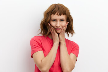photo of a cute, thoughtful red-haired woman in a red T-shirt standing on a background and holding her hands near her face. Studio photo with an empty space for inserting an advertising layout