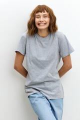 Obraz premium a wide-smiling, joyful woman stands in a gray T-shirt on a white background, leaning against the wall. Vertical Studio Photography