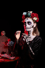 Young adult wearing dios de los muertos body art to look like traditional cartina skull in studio, having flowers crown and santa muerte costume. Mexican holiday tradition on halloween day.