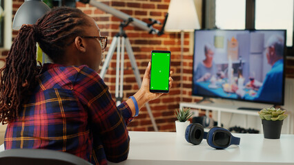 Fototapeta na wymiar African american adult vertically holding smartphone with greenscreen, sitting at home desk. Working with blank copy space mock up template and isolated chroma key display. Tripod shot.