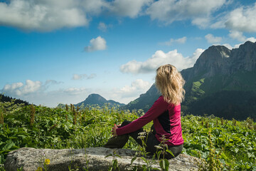 Naklejka premium A tourist girl in a purple sweater sits on a large rock against a mountain backdrop. Side view. High grass, blue sky with clouds. Travel and tourism. Mountain hike.