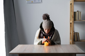 Woman freezing at home, sitting at the table warming hands of burning candle. Woman with home...