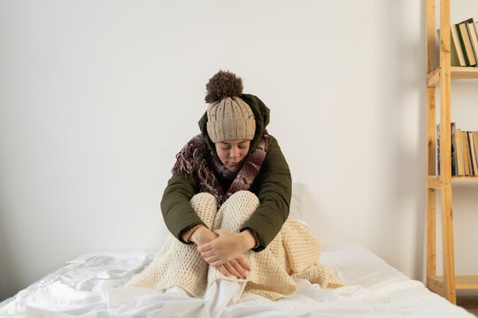 Angry young woman warmly dressed in a cold house sits on the bed feeling cold inside the house