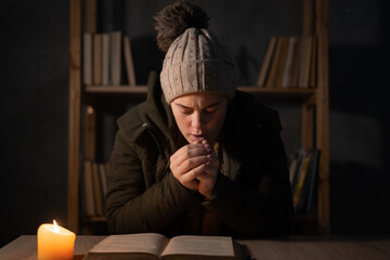 Freezing young woman in winter clothes warms her hands on lights with candles. Shutdown of heating...
