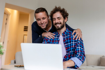 Lovely gay couple laughing together while sitting in their living room at home. Two romantic young male lovers having fun surfing the internet indoors. Young gay couple living together.