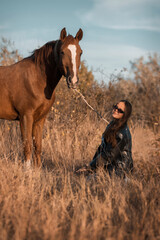 A white girl in leather jacket, black glasses next to a horse and poses in the forest at sunset, like in a fairy tale. Well-groomed face with beautiful cosmetics. Enjoy nature and animals. Horse