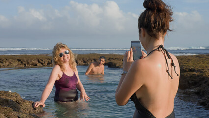 Happy family on holiday at ocean shore. Adult woman taking picture of senior woman as they are standing in sea water inside reef pool and man swimming towards waves. Happy time on wild beach in summer