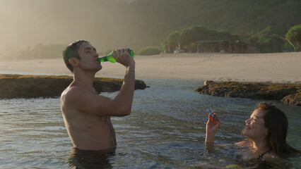 A happy couple bathing in natural ocean reef pool sharing a bottle of beer. Woman giving her husband a refreshing alcoholic drink as she is swimming with cellphone at sunset on beach. Soft focus.