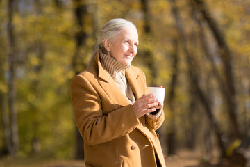 elderly woman with gray hair  in yellow coat drinks tea in autumn forest. copy space. mental health. Slow life. Enjoying the little things. nature walks.
