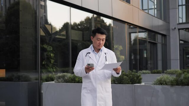 Adult male asian doctor walking holding document, texting message, scrolling screen of mobile phone. Middle age doctor sends an online prescription to the patient viral infection, outside clinic.
