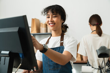 Happy young Asian woman cashier wears an apron and using pos terminal to input orders on coffee...