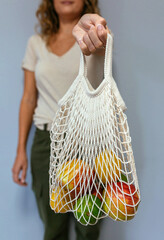 Unrecognizable woman holding reusable mesh shopping bag with fruits inside over grey background. Sustainable lifestyle and zero waste concept.
