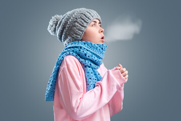 Young woman in a knitted hat and blue scarf is trying to keep warm, breathing. Gray background. The...