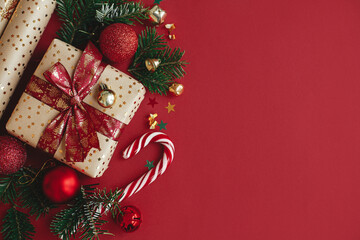 Modern christmas flat lay. Stylish christmas gift, wrapping paper,  fir branches, candy cane and baubles composition on red background, copy space. Merry Christmas and Happy Holidays!