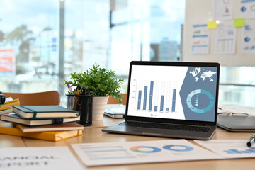Front view of laptop with financial graph data on screen and office supplies on wooden office desk